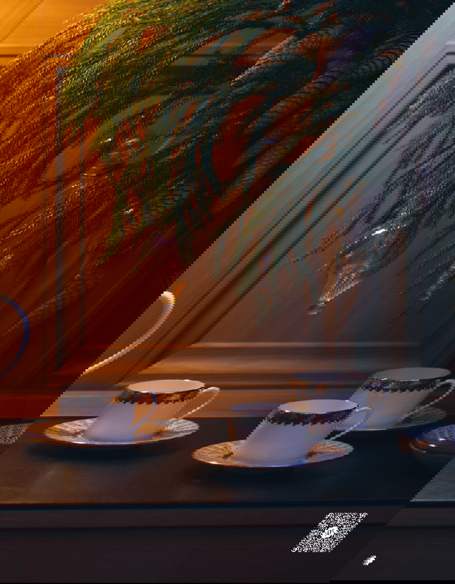 Крупный план чашек и блюдец на тёмной поверхности, над ними висят еловые ветви и стеклянные ёлочные украшения в съёмке интерьера.