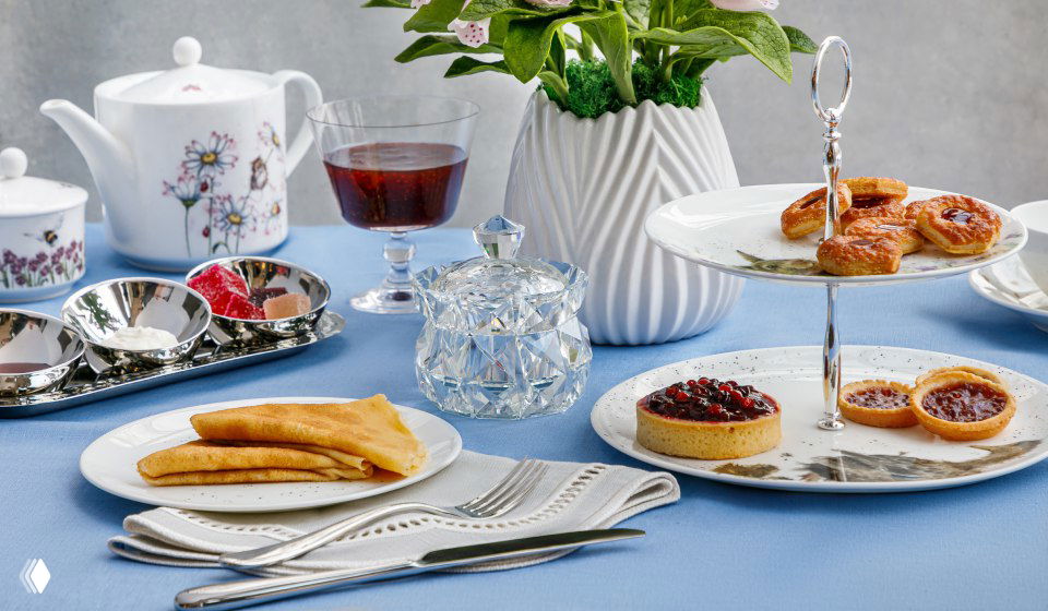 Детальная подача на голубой скатерти: чайник, этажерка с пирожными, хрустальная сахарница и тарелка с блинами