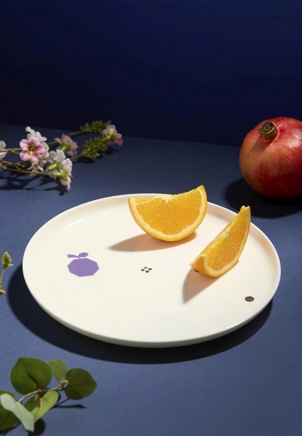 Минималистичная тарелка из коллекции «Волшебные фрукты» на синем фоне: белый фарфор, фиолетовый фруктовый мотив и ломтики апельсина.
