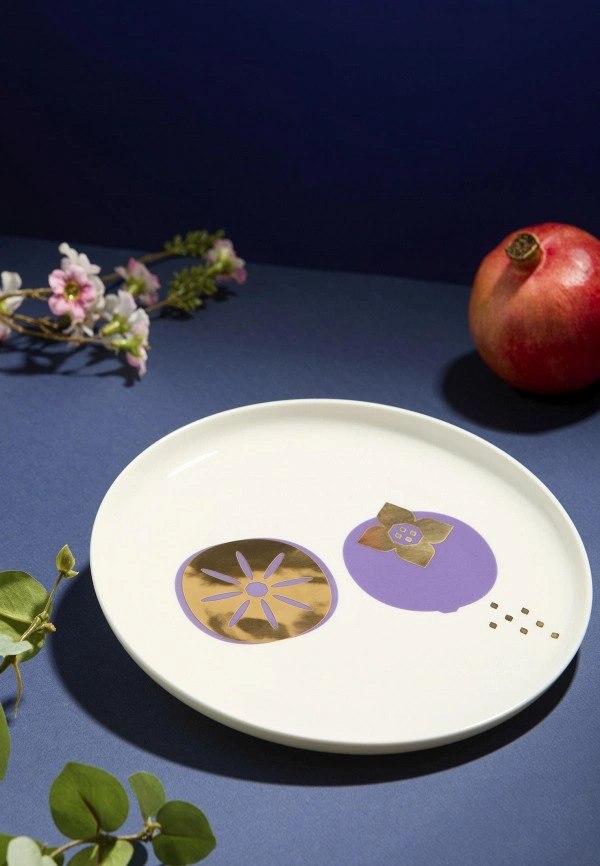 Натюрморт с тарелкой коллекции «Волшебные фрукты»: графические плоды в фиолетовом и золотом на белом костяном фарфоре и гранат рядом.