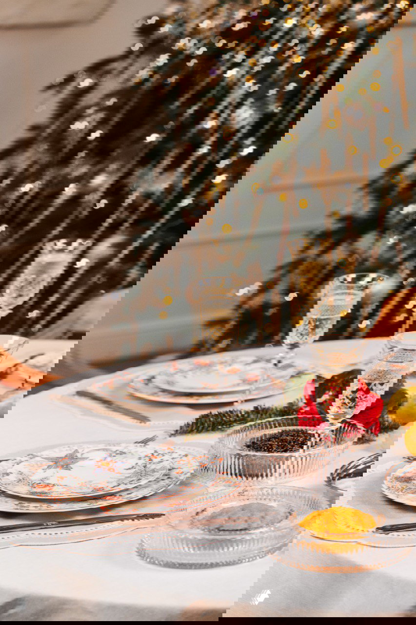 Горизонтальное фото: праздничный стол с сервировкой из коллекции «Счастливая звезда», бокалами и размытой ёлкой на заднем плане для атмосферного кадра.