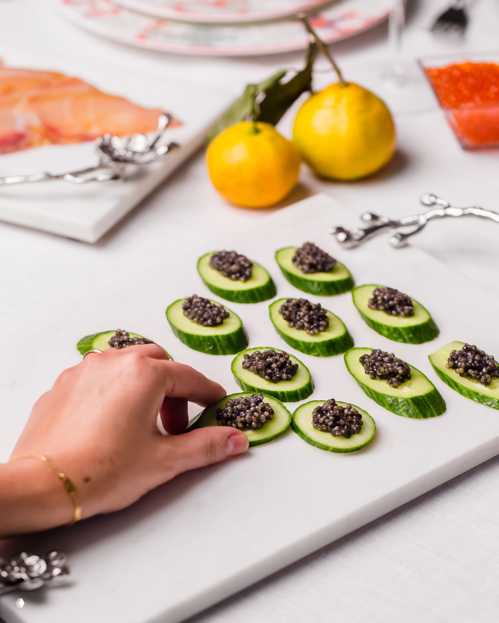 Канапе из ломтиков огурца с порциями черной икры на мраморной доске, рядом фрукты и столовые аксессуары — вид в композиции для закусок.