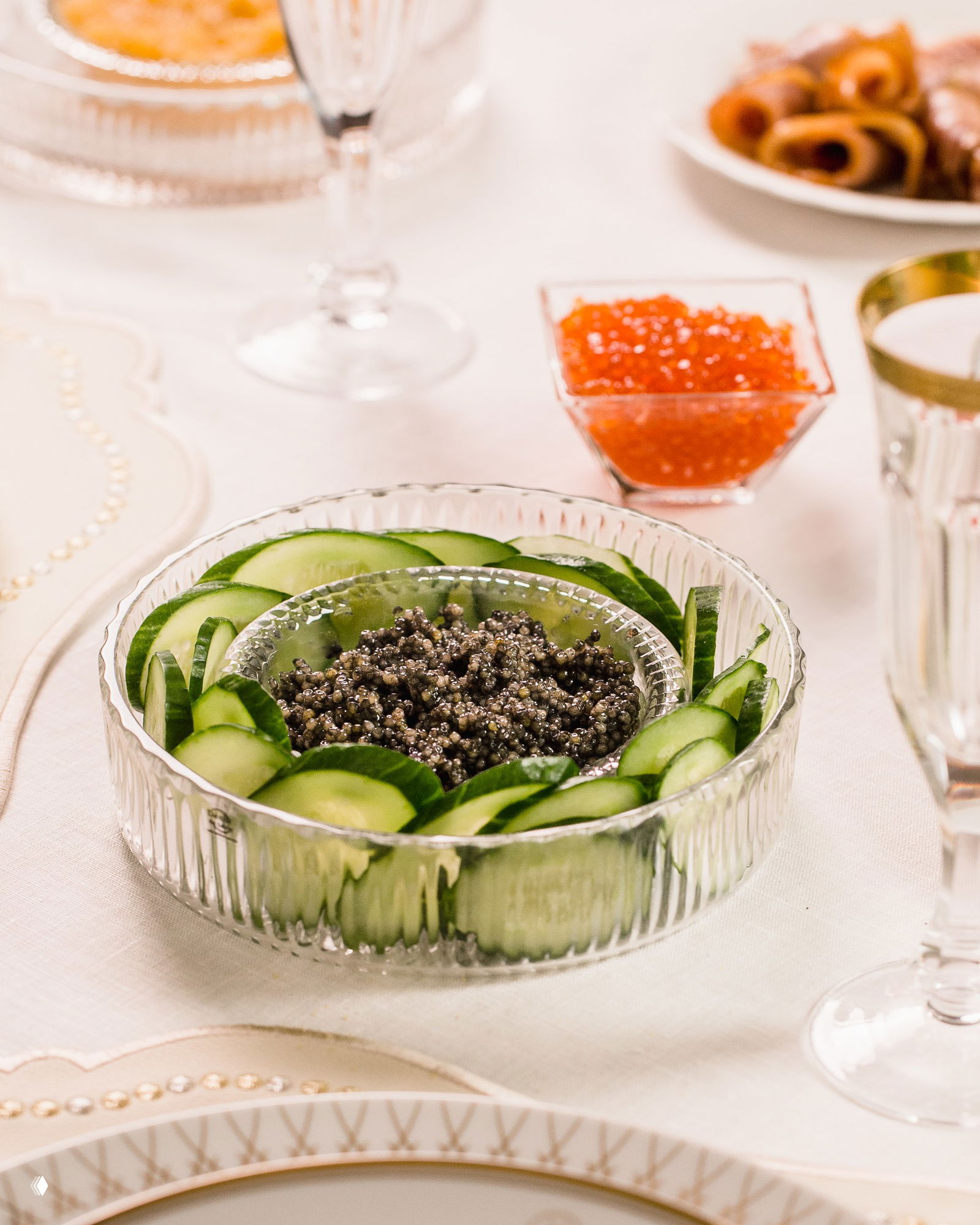 Икорница с черной икрой окружена тонко нарезанными ломтиками огурца и бокалом шампанского на фоне елочных огней — стильный кадр сервировки.