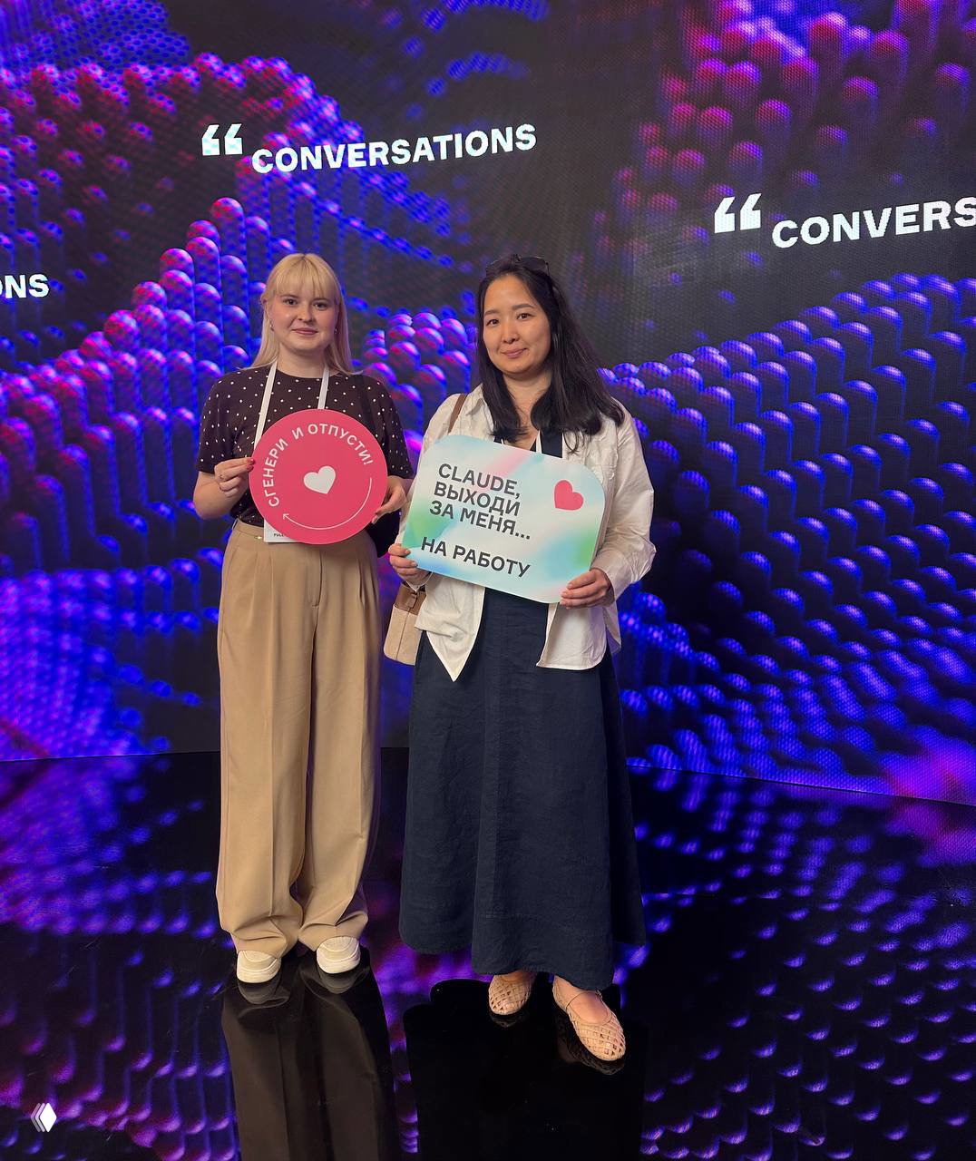Две участницы на фоне светодиодного экрана «CONVERSATIONS», улыбаются и держат таблички с надписями о генераторах и Claude на мероприятии по AI.