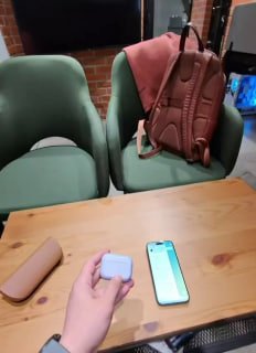 Рука держит кейс наушников на столе рядом со смартфоном и рюкзаком — кадр из тестирования гаджетов в кафе.