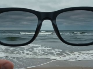 Черный каркас очков Wayfarer на переднем плане; через линзы виден прибрежный пейзаж с морем, волнами и пасмурным небом, фон размытый.