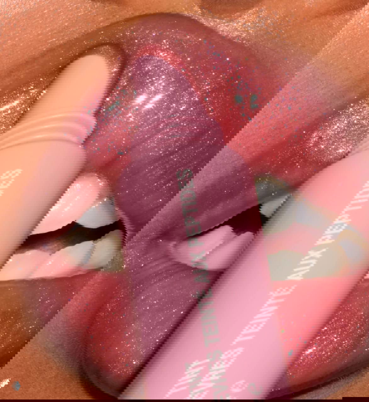 Макрокадр губ с блестящим розовым тинтом Rhode; тюбик на переднем плане показывает текстуру и шиммер продукта.