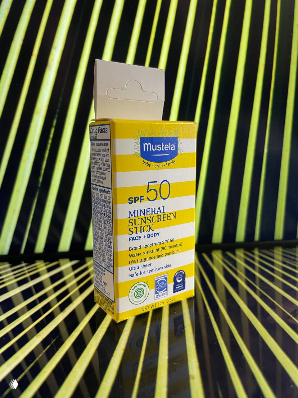 Коробка солнцезащитного стика Mustela SPF50 на фоне с диагональными жёлто‑чёрными полосами; вид спереди с акцентом на бренд и SPF.