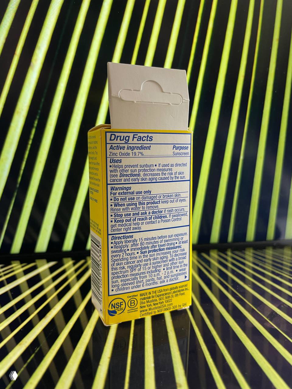 Задняя сторона упаковки Mustela с табличкой Drug Facts и инструкцией на английском языке; коробка размещена на полосатом фоне.