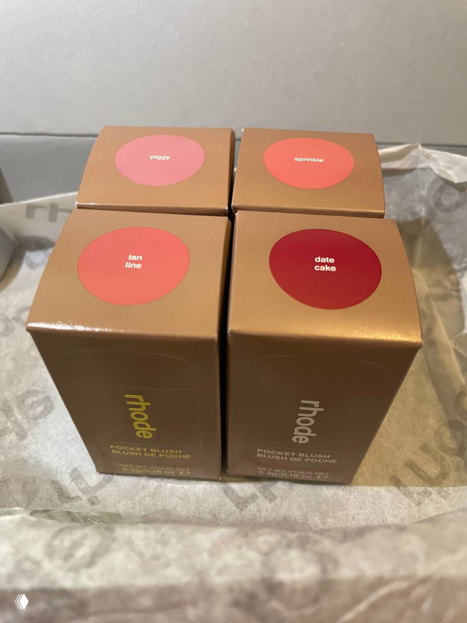 Четыре коробочки румян Rhode в плотной упаковке с круглыми наклейками оттенков (piggy, sprinkle, tan line, date cake) на фоне бумаги из посылки.