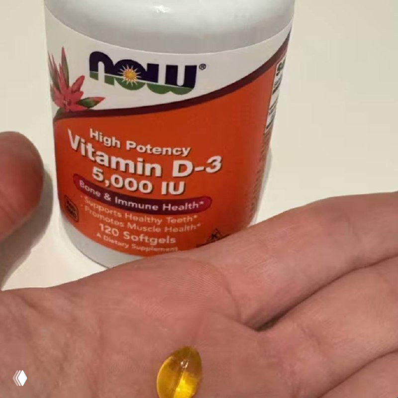 Бутылка Vitamin D-3 5000 IU (NOW) и желтая мягкая капсула на ладони — демонстрация формы и размера софтагела витамина D.