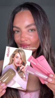 Модель демонстрирует несколько тюбиков блесков Rhode и промо‑лист с изображением, кадр фокусируется на лице и упаковке продукта