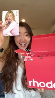 Радостная девушка распаковывает ярко‑розовую коробку Rhode, видно внутренние пакеты и первые образцы свотчей блесков