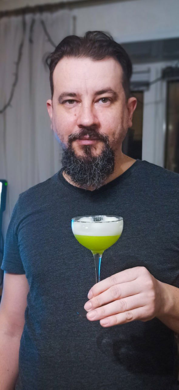 Человек с бородой держит в руке бокал с зелёным коктейлем «Lucky clover» с белой пеной; видна верхняя часть туловища и фон кухни