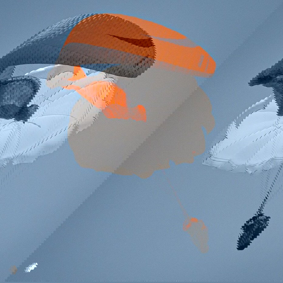 Резервный круглый парашют полностью раскрыт под основным оранжевым крылом, пилот в подвеске, видно купол и стропы на фоне неба.