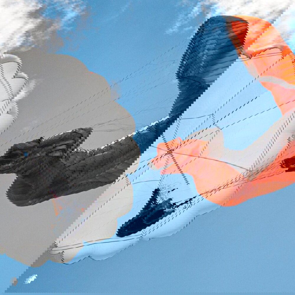 Крупный план раскрытого белого запасного купола и складывающегося оранжевого основного крыла, стропы натянуты, небо с облаками.