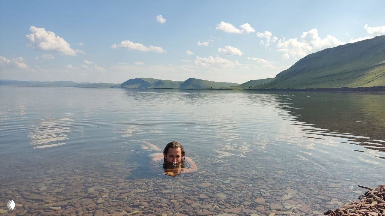 Человек купается в прозрачной воде Красноярского водохранилища у галечного берега на фоне гористого ландшафта и спокойного неба.