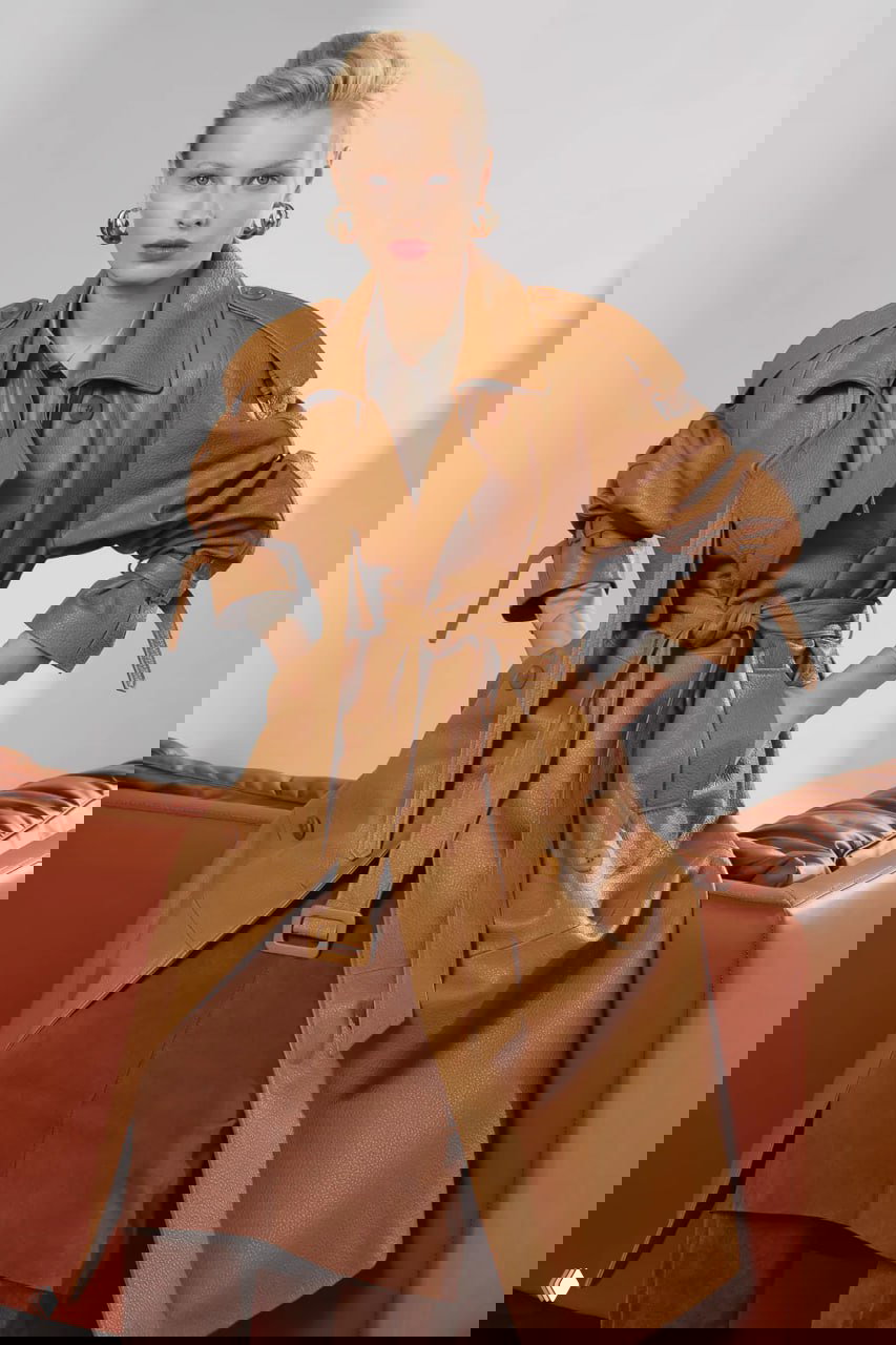 Фронтальный ракурс модели в кожаном тренче с поясом и объемными рукавами, акцент на текстуре кожи и посадке изделия.