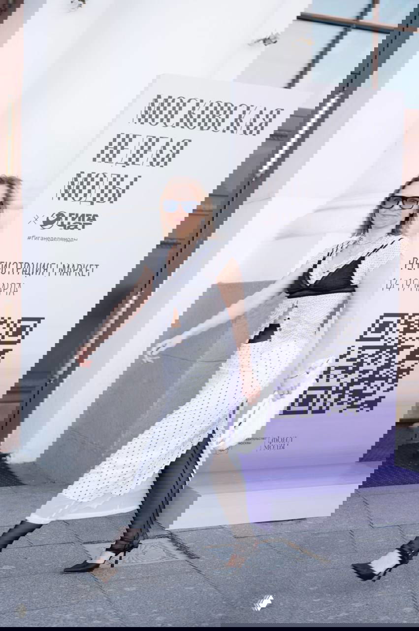 Прогулочный кадр у баннера «Московская неделя моды»: белое платье в горох с чёрным корсетом, чёрные колготки и туфли на каблуке, образ для стрит‑стайла.