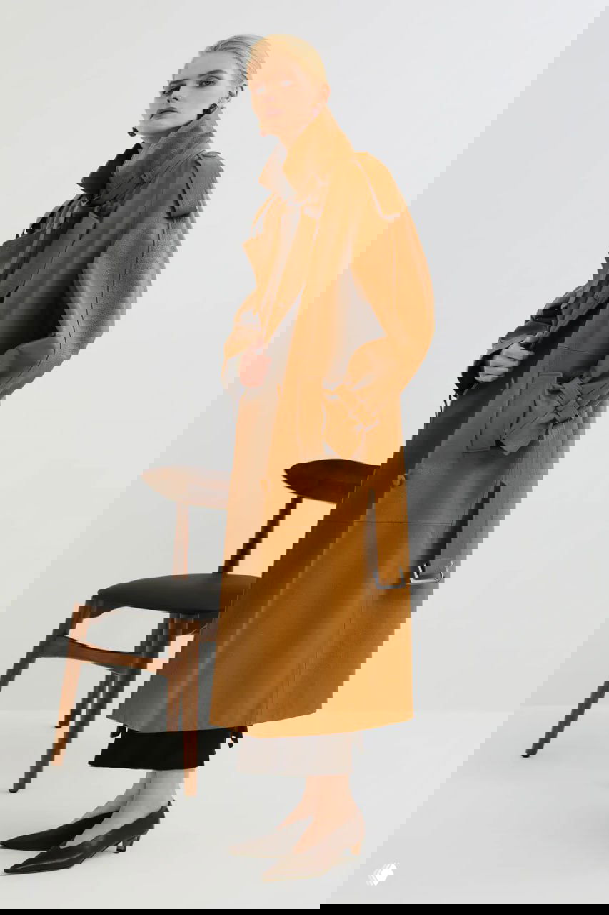 Боковой профиль модели в карамельном тренче на минималистичном фоне, демонстрирующий длину, разрезы и силуэт пальто.