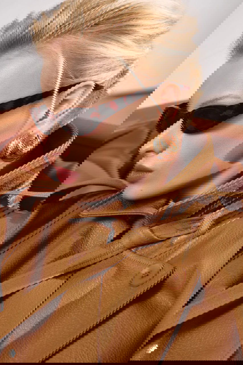 Крупный план воротника и декоративных деталей кожаного тренча, аксессуары — солнцезащитные очки и золотистые серьги на модели.