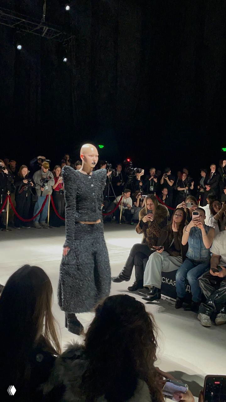 Андрогинная модель в текстурированном чёрном костюме на показе Capparel: публика с телефонами и пресс на заднем плане.