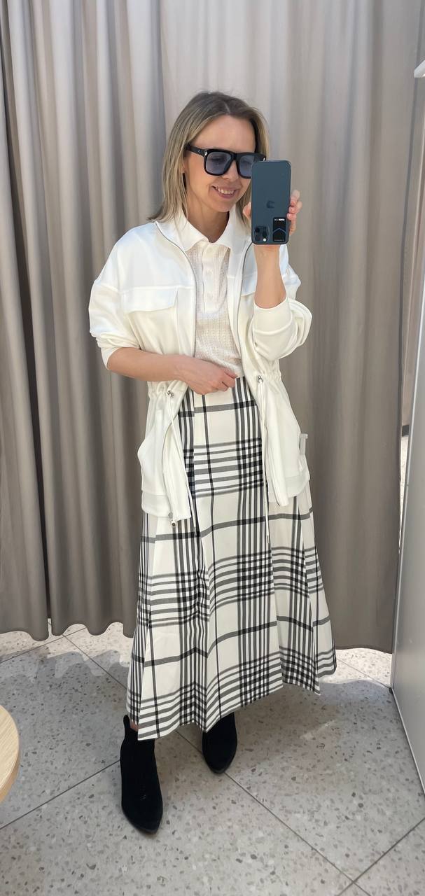 Модель в белой парке-куртке поверх светлого пуловера и клетчатой юбки, образ дополнен тёмными солнцезащитными очками и ботильонами.