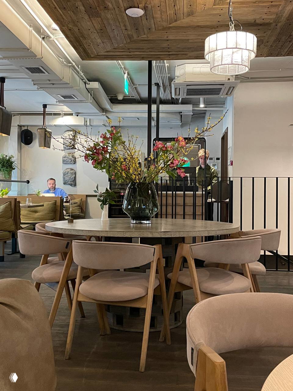 Интерьер кафе М2 в ЗИЛ Арт: круглый деревянный стол с большой вазой свежих цветов, розово‑бежевые мягкие стулья, лампы, деревянный потолок и уютная атмосфера.