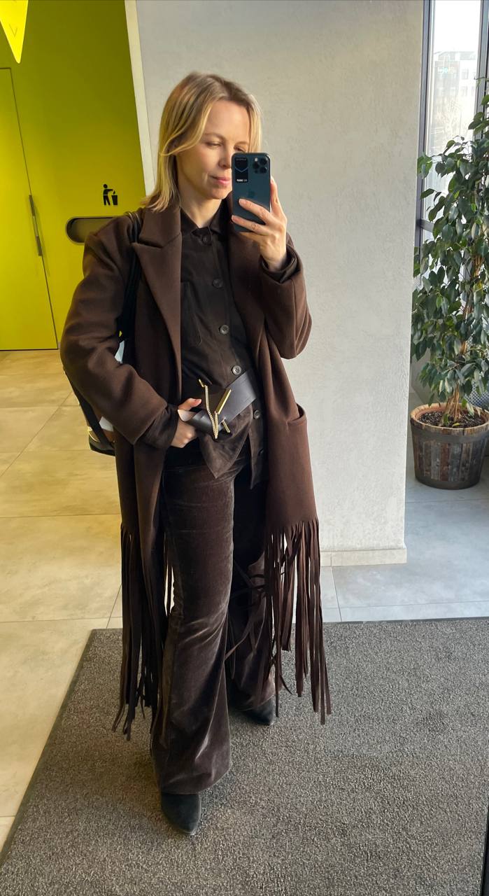 Зеркальное фото в коричневом пальто с бахромой и бархатных широких брюках; сидящая растительность и входная зона на заднем плане.