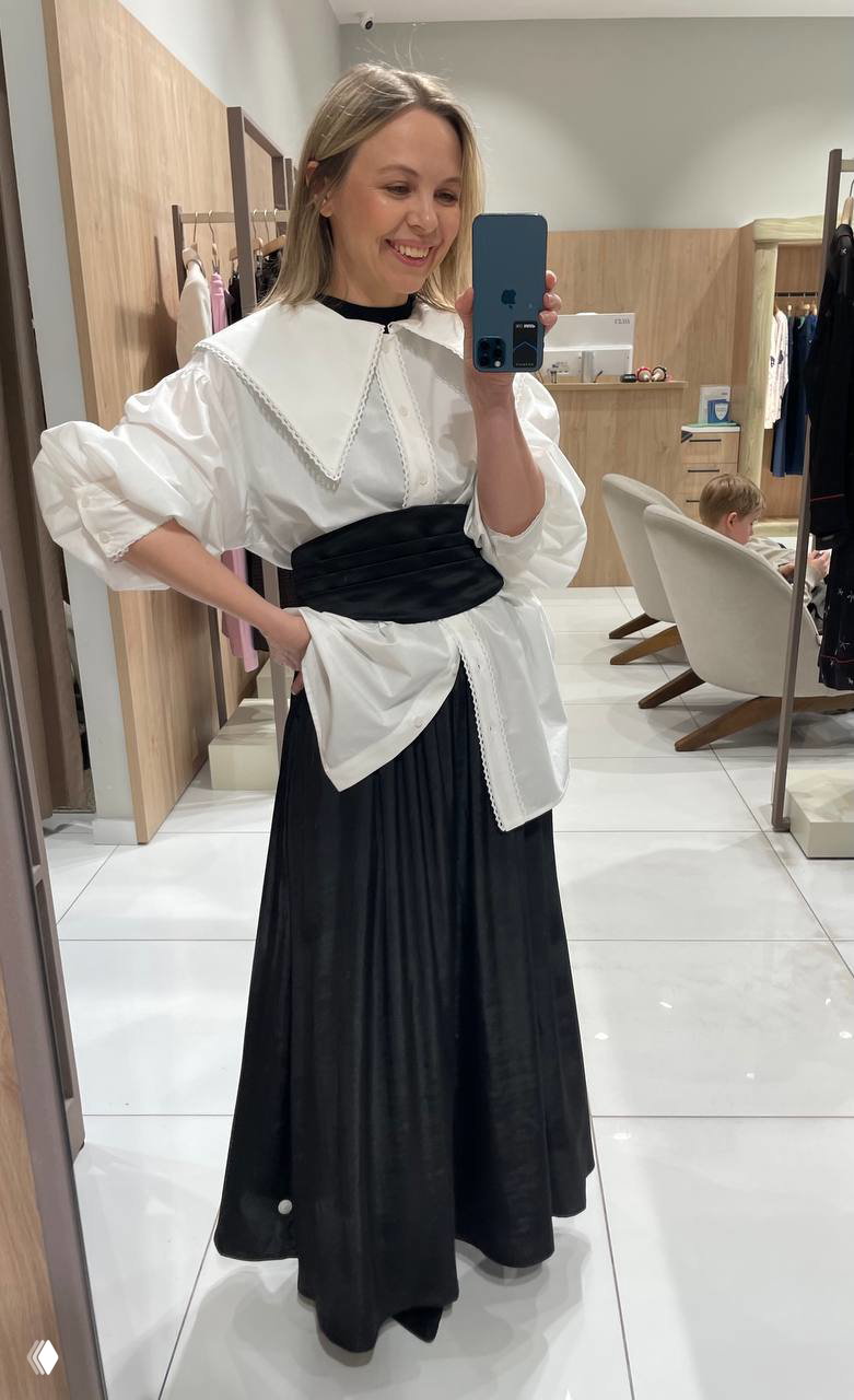 Селфи в зеркале: белая блуза с крупным воротником и чёрная юбка, примерка в бутике, на заднем плане стойки с одеждой и аксессуарами.