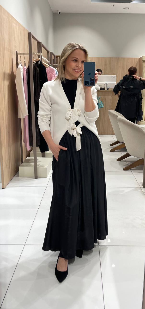 Женщина в зеркале примерочной в белом кардигане и чёрной юбке, улыбается, видна мебель бутика и другие покупатели на заднем фоне.