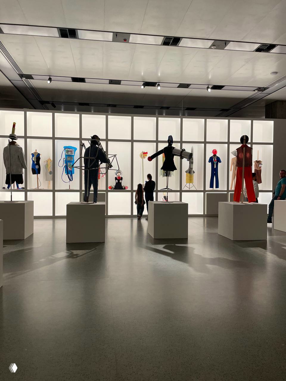 Выставочная секция с манекенами: ассорти костюмов и арт‑объектов на белых пьедесталах на фоне ярко подсвеченной задней стены, посетители рассматривают экспонаты.