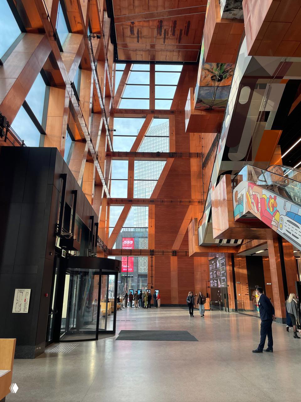 Вестибюль ЗИЛ Арт: высокий стеклянно‑деревянный атриум с крупными медными балками, панорамными окнами, эскалатором и прохожими у входа, видна городская улица.