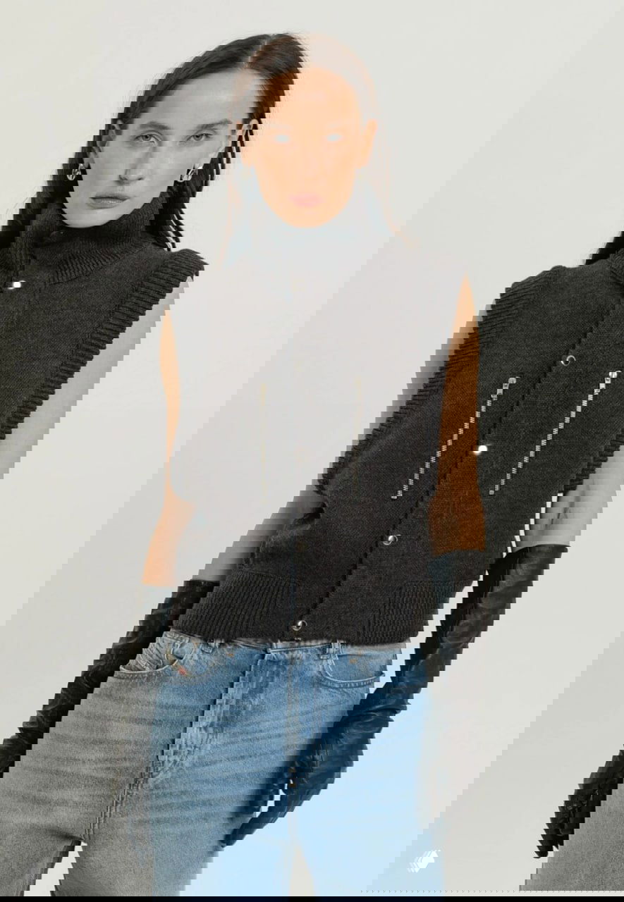 Модель в тёмном вязаном безрукавном жилете с высокой горловиной и декоративными молниями, образ с джинсами и длинными кожаными перчатками.