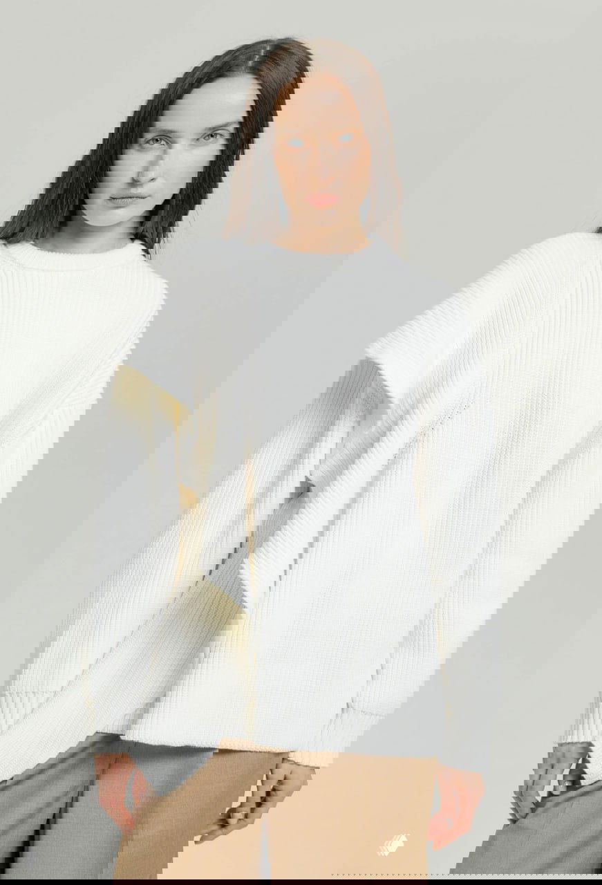 Модель в белом вязаном свитере с асимметричным драпированием, романтичный и мягкий образ на светлом фоне, крупный план по талии.