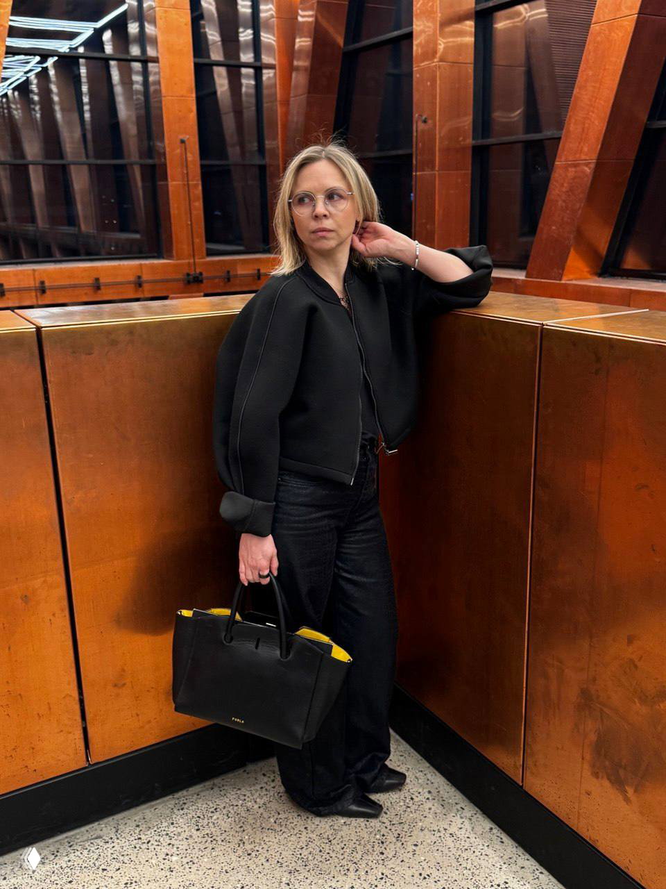 Портрет в интерьере: автор блога в чёрной куртке и широких брюках с чёрной сумкой с ярко‑жёлтой подкладкой позирует у медной панели и больших окон внутри ЗИЛ Арт.