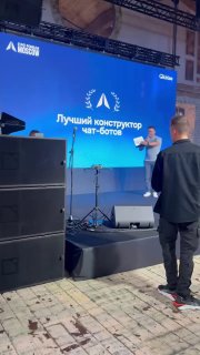 Вертикальная фотография сцены: спикер у микрофона на фоне синего экрана с текстом о победе puzzlebot в номинации «Лучший конструктор чат-ботов».