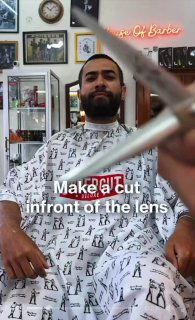 Клиент в барбершопе в накидке для стрижки, барбер держит ножницы крупным планом перед объективом; видно интерьер, полки и неоновую вывеску.