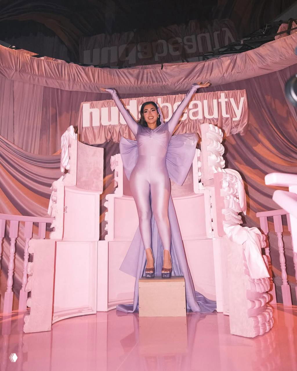 Фотография с мероприятия: женщина в облегающем сиреневом костюме стоит на сцене перед баннером бренда и праздничной декорацией, выражение торжества.