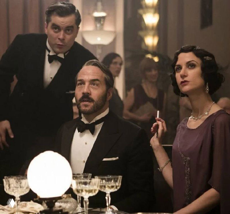 Мужчина и женщина в вечерней одежде сидят за столом с бокалами и лампой в интерьере начала XX века — сцена исторической драмы.