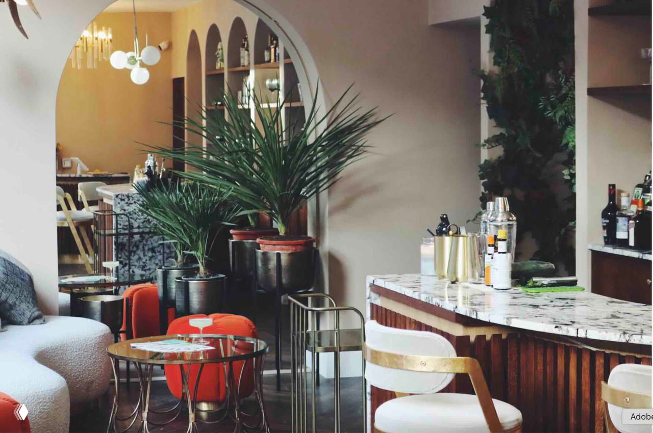 Интерьер комбинированного места: барная стойка, высокие стулья, декоративные растения и мягкая зона ожидания — пример оформления салона с баром.