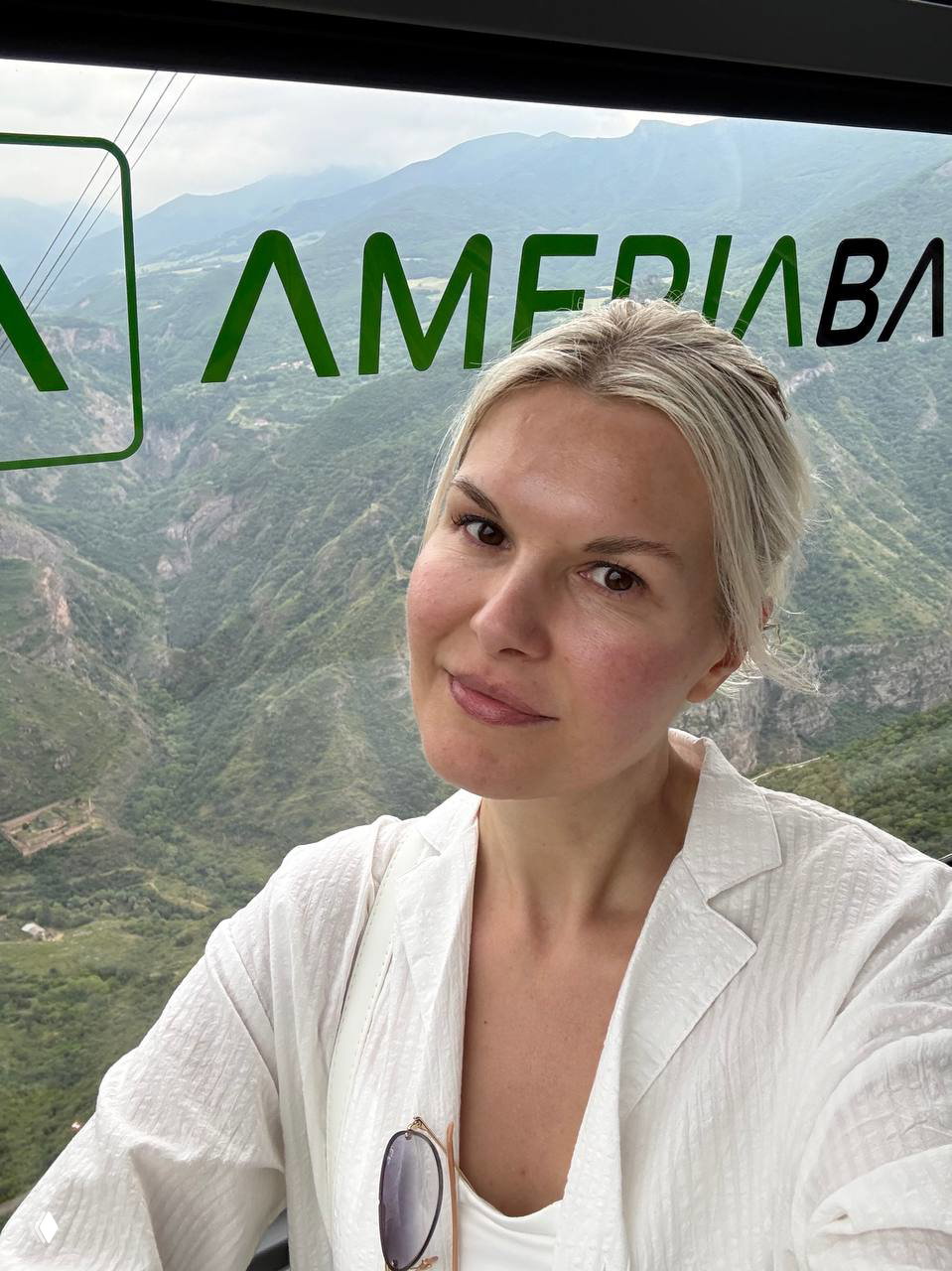 Селфи женщины в кабинке канатной дороги на фоне глубоких зелёных ущелий, на стекле виден логотип банка и виды горного ландшафта.