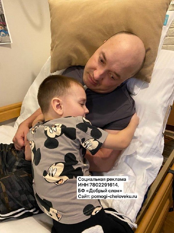 Мужчина в больничной постели обнимает маленького ребёнка; на кадре видна наклейка с данными благотворительного фонда и ссылкой для помощи семьям.