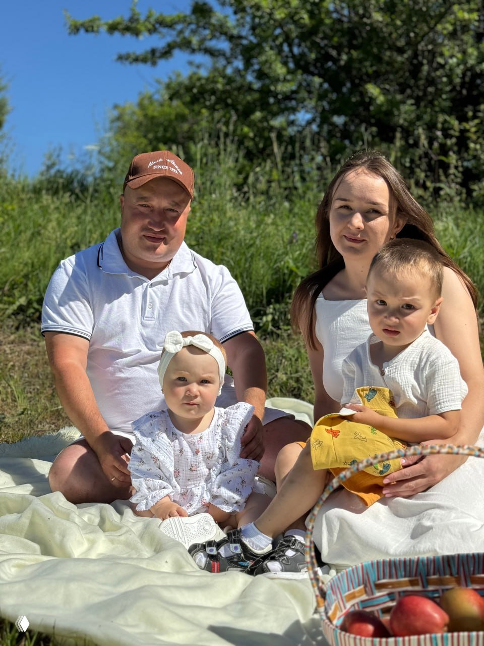 Семейная летняя фотография на природе: мужчина, женщина и двое маленьких детей сидят на пледе среди зелёной травы; пикник с корзиной фруктов.