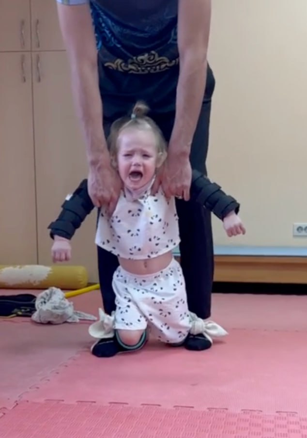 Девочка в фиксирующих повязках и корсете стоит с поддержкой взрослого на реабилитации в зале; упражнение вызывает слёзы и усилия.