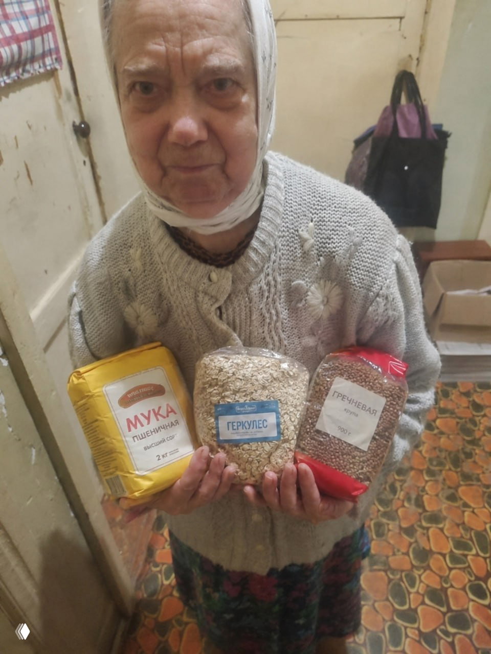 Пожилая женщина в коридоре держит пакеты с мукой, геркулесом и гречкой — волонтёрская помощь для сельского пенсионера.