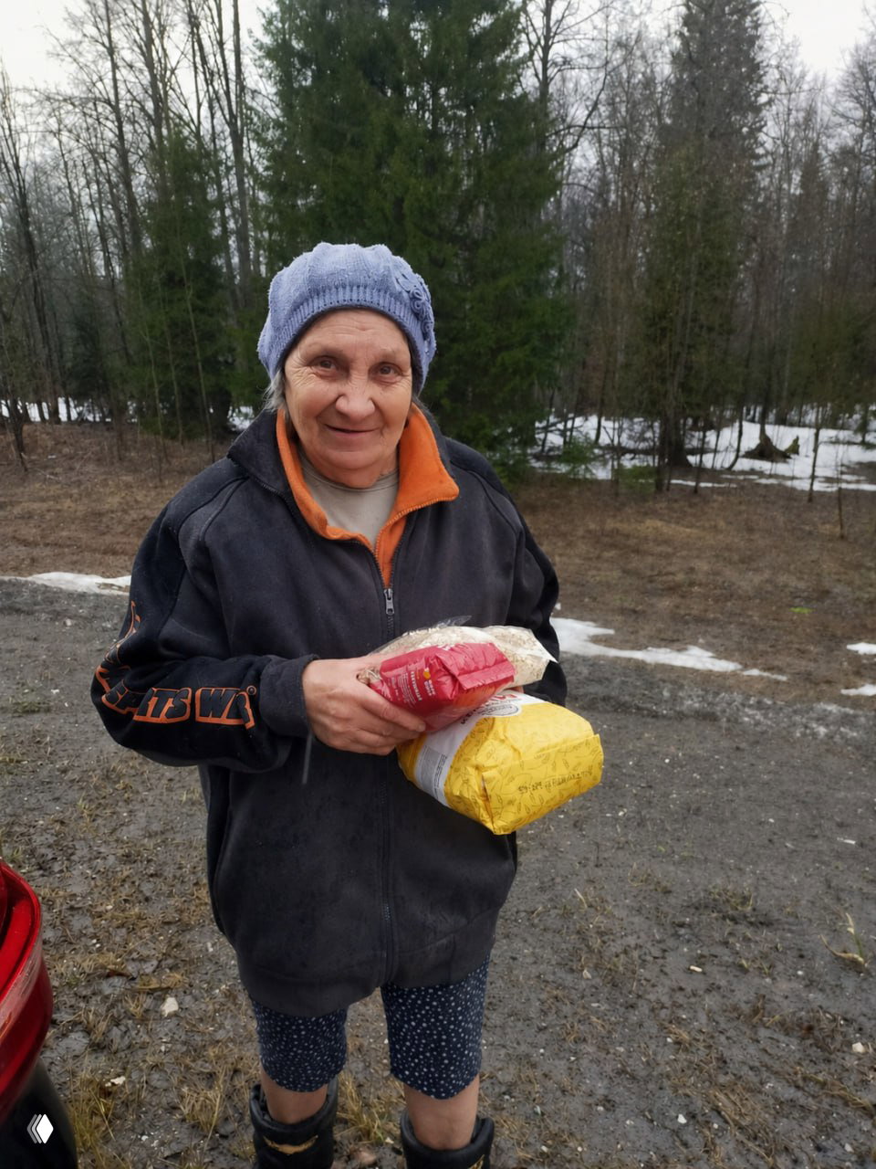 Улыбающаяся женщина на фоне леса держит пакеты с продуктами; фото демонстрирует доставку питания волонтёрами в деревню.
