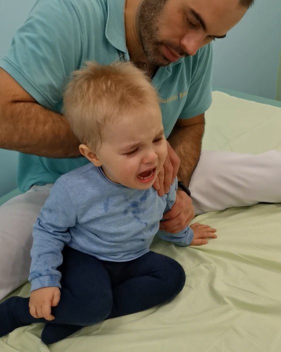 Маленький ребёнок плачет во время реабилитационного занятия; видно терапевта, поддерживающего и помогая выполнять упражнения.