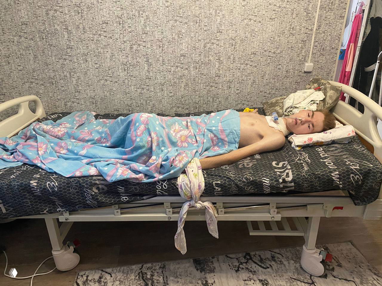 Лежащий в кровати молодой человек с видимой дыхательной трубкой, покрытый пледом; домашняя или палатная обстановка во время продолжительной реабилитации после травмы.