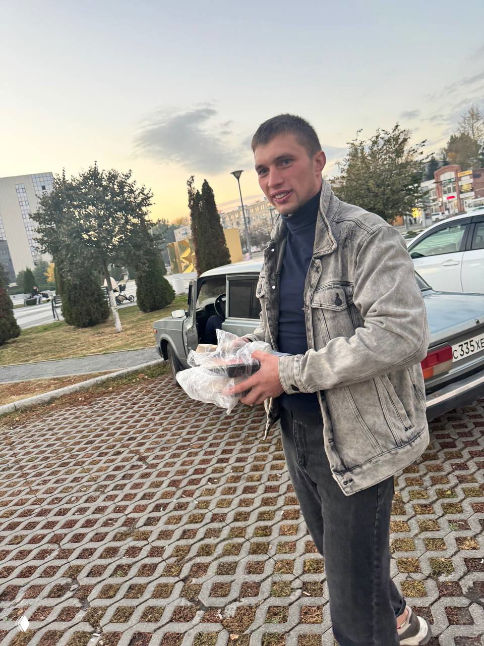 Молодой мужчина улыбается на улице у автомобиля с пакетами в руках; кадр сделан до травмы, видно обычную городскую обстановку и спокойный фон.